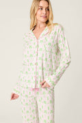 Wearing the P.J. Salvage Playful Prints Island Time Long Sleeve Top, a woman stands against a plain light background. The white cotton-modal pajama features light green palm trees and pink pineapples, with buttons and a relaxed fit.