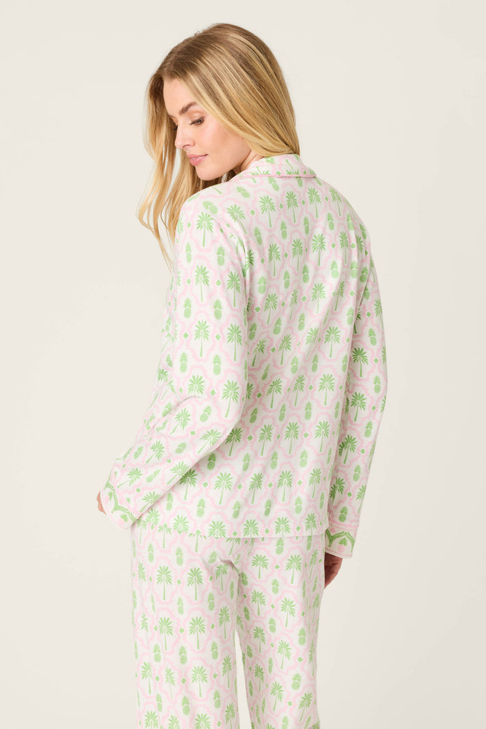A woman with long blonde hair models the P.J. Salvage Playful Prints Island Time Long Sleeve Top, featuring a light background with green palm tree print, as she stands mostly with her back to the camera, looking over her shoulder.