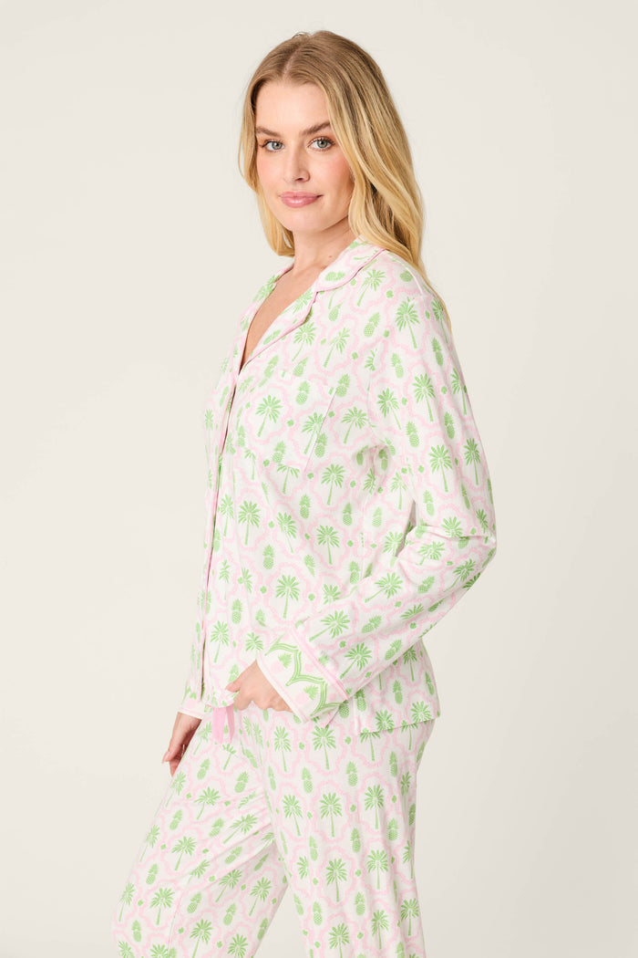 A woman with long blonde hair poses sideways in the P.J. Salvage Playful Prints Island Time Long Sleeve Top, featuring a green palm tree pattern with pink accents on soft cotton-modal jersey, set against a plain light background.