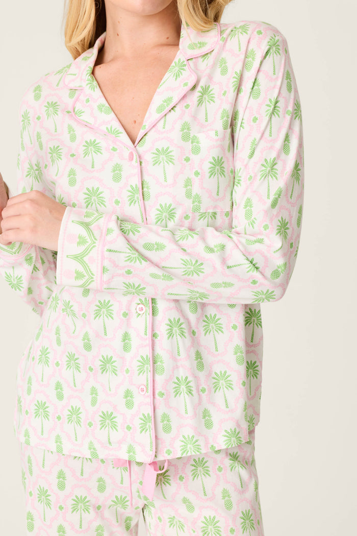 A person wears the Playful Prints Island Time Long Sleeve Top by P.J. Salvage, featuring an ivory base with a light pink and green palm tree and pineapple print. The collared, buttoned pajama shirt is shown on the upper body only.