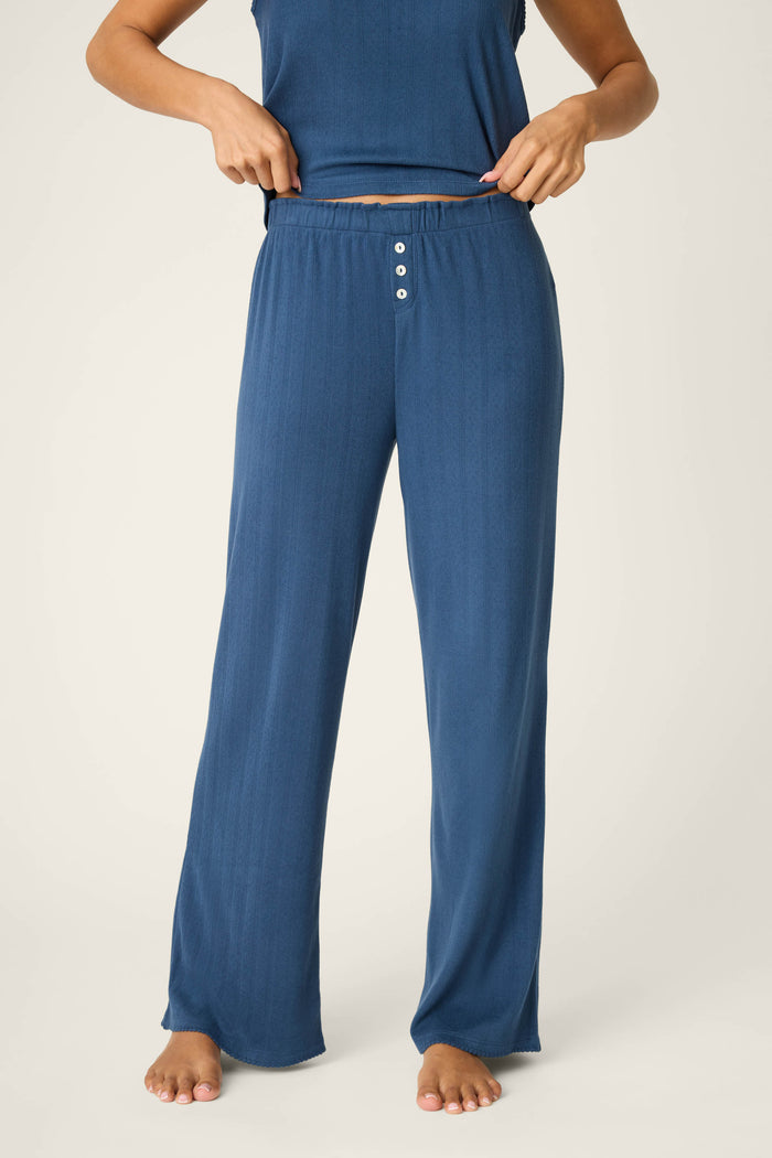 A person stands barefoot adjusting the waistband of P.J. Salvage Pointelle Essentials Deep Sea Open Leg Pants—loose-fitting, high-waisted lounge pants in blue with an adjustable drawstring and three front buttons, paired with a matching sleeveless top.