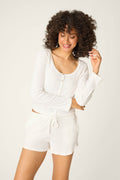 A person with curly hair smiles while wearing the P.J. Salvage Pointelle Essentials Ivory Cardigan, paired with matching white shorts, standing against a plain, light background.
