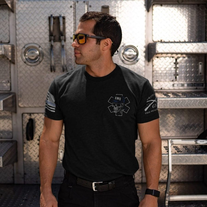 A man wearing a black short-sleeve tactical-style T-shirt with white EMS and tactical insignia on the chest and sleeves, standing in front of a metallic diamond-plate vehicle door.