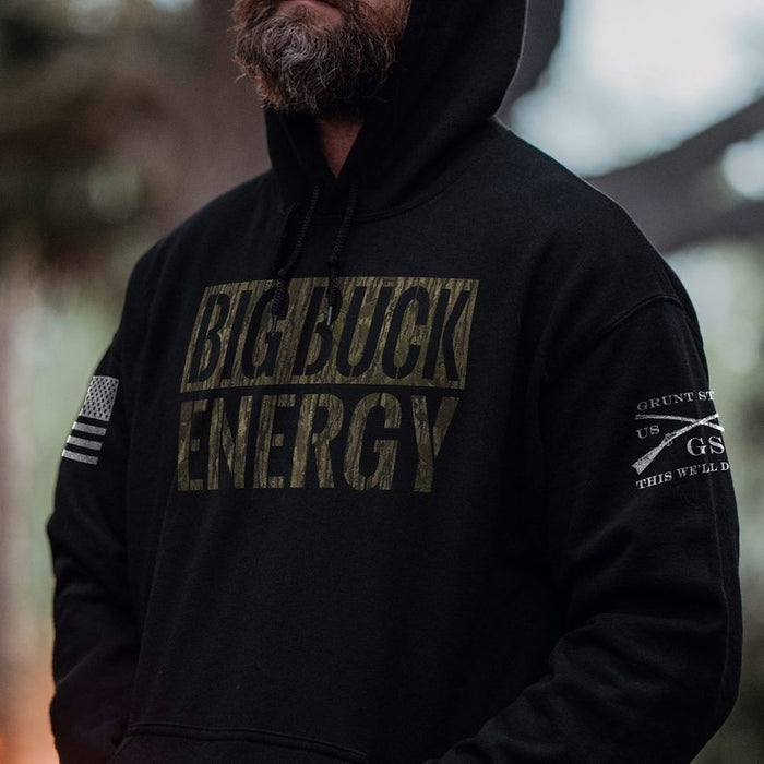 A man wearing a black pullover hoodie with a large distressed 'BIG BUCK ENERGY' logo printed across the chest in a muted olive-brown camouflage pattern. The hoodie also features a small American flag patch on the right sleeve and a white graphic on the left sleeve.