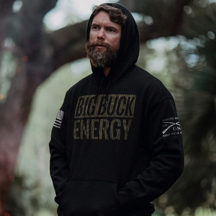 A man with a beard wearing a black hoodie with distressed 'BIG BUCK ENERGY' text on the chest, standing outdoors in a wooded setting.