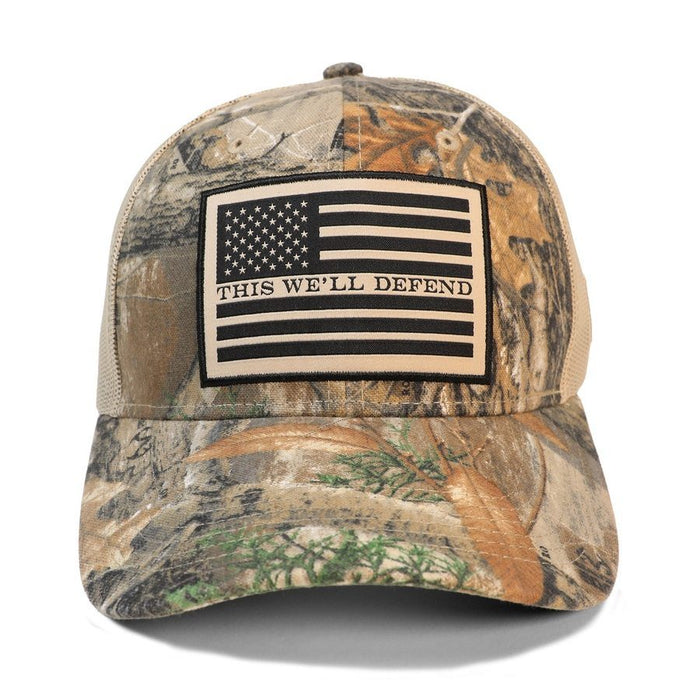 A camouflage trucker hat with a beige mesh back and a front patch featuring a black and beige American flag with the text 'THIS WE'LL DEFEND'.