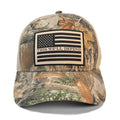 A camouflage trucker hat with a beige mesh back and a front patch featuring a black and beige American flag with the text 'THIS WE'LL DEFEND'.