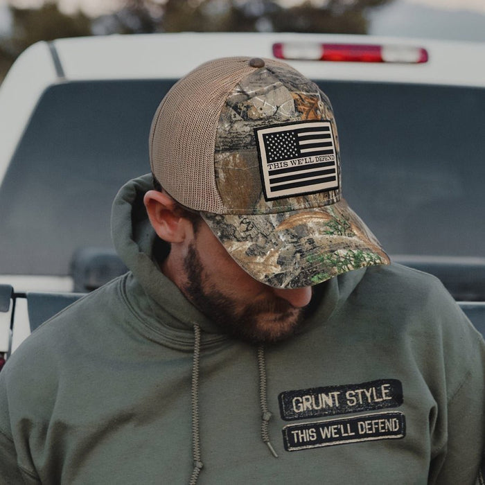 A man wearing a camouflage trucker hat with a US flag patch that reads 'GRUNT STYLE THIS WE'LL DEFEND' and a matching olive green hoodie with a matching chest patch, standing in front of a white vehicle.