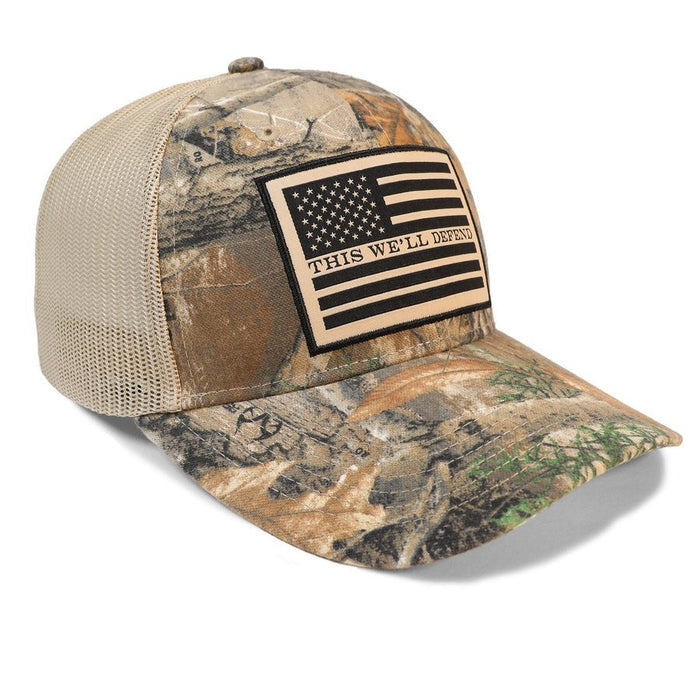 A camouflage trucker hat with a beige mesh back and a black-and-beige American flag patch on the front that reads 'THIS WE'LL DEFEND'.