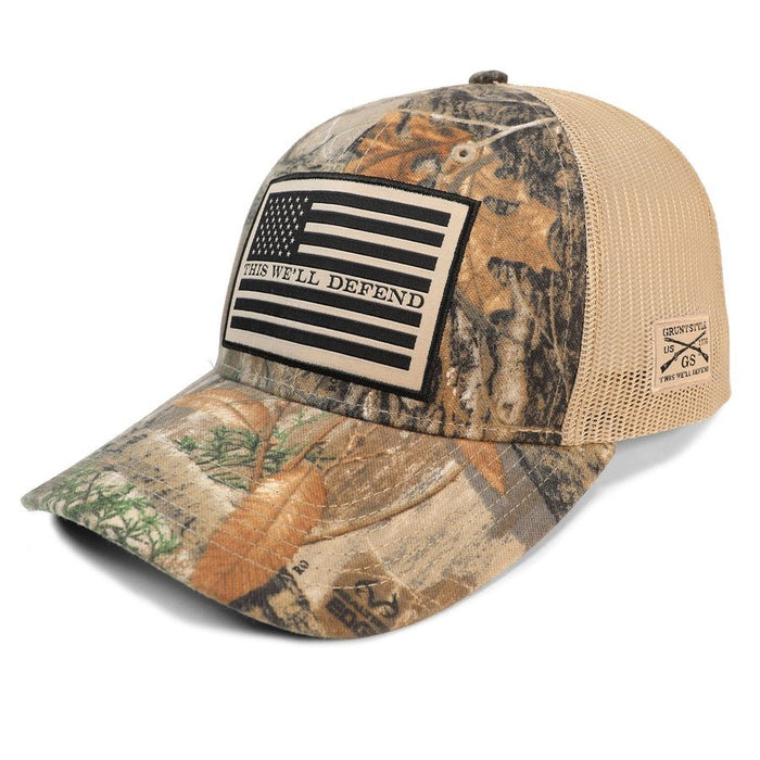 A camouflage trucker hat with a tan mesh back, featuring a black and beige 'This We'll Defend' American flag patch on the front and a small 'Spartan Outdoors' logo on the side.