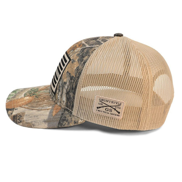 A trucker-style baseball cap with a tan mesh back and a camouflage patterned front panel and brim. The back features a tan mesh back with a small rectangular brand patch that reads 'Grunt Style 1975 GS Trucker System'.