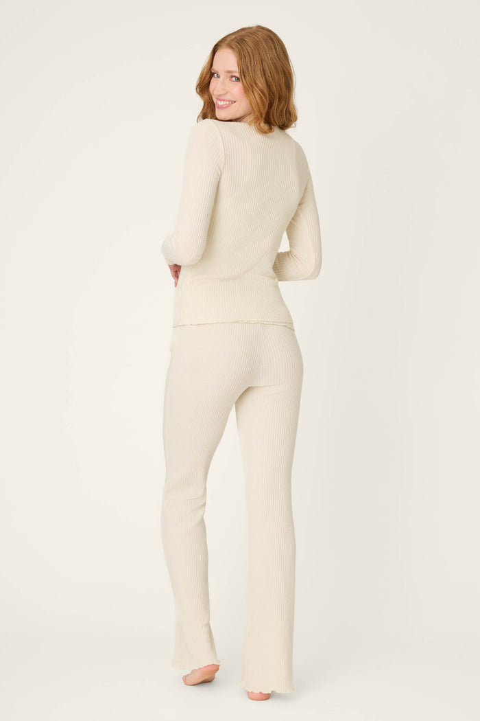 A person with long, wavy, auburn hair stands barefoot, facing away from the camera in the P.J. Salvage Reloved Rib Natural 3-Piece Lounge Set made with REPREVE yarn, against an off-white background.