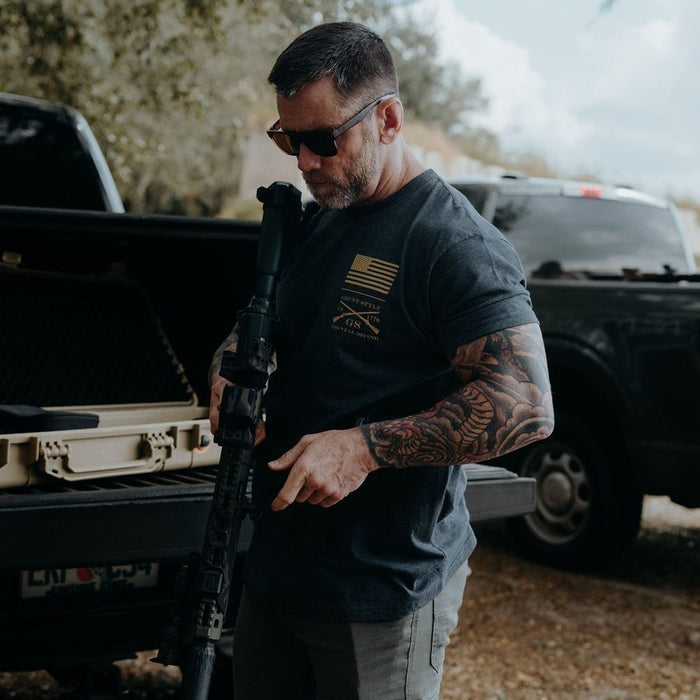 Man with tattoos holding a rifle in front of a truck