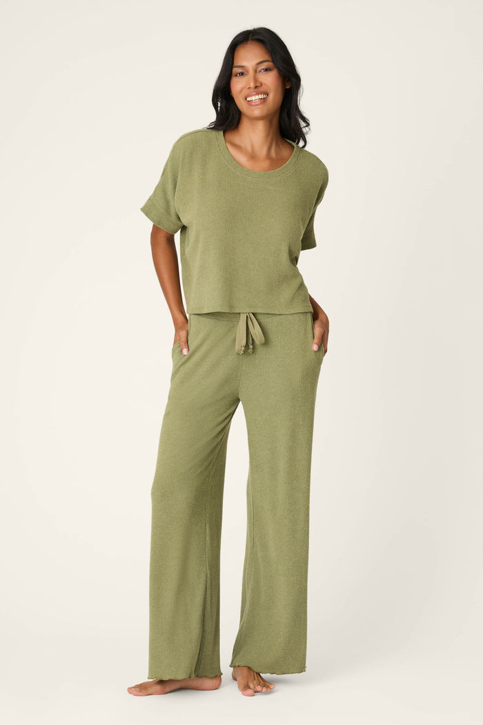 A woman smiles barefoot in the Olive You Forever Lounge Set by P.J. Salvage—an olive green, loose-fitting short-sleeve top with matching wide-leg, drawstring pants—posed against a plain light background.