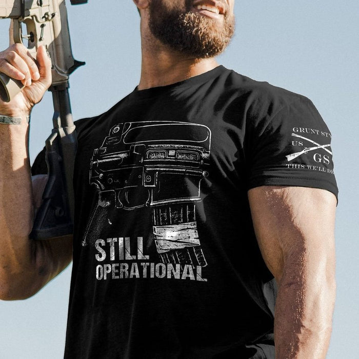 A close-up lifestyle shot of a man wearing a black t-shirt with a white graphic of a rifle magazine and the text 'STILL OPERATIONAL'. He is holding a rifle against a clear blue sky background.