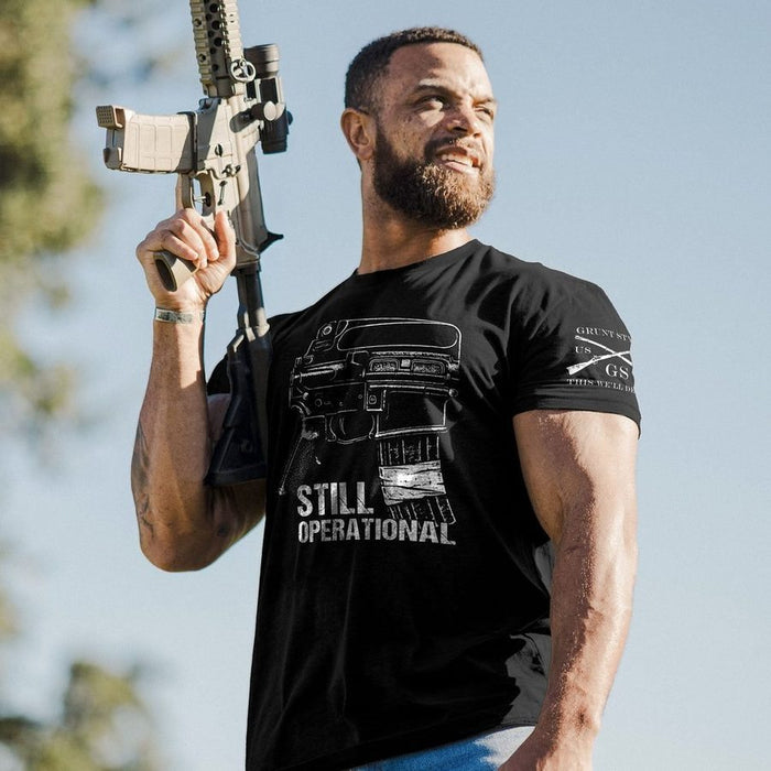 A muscular man outdoors wearing a black graphic t-shirt that reads 'STILL OPERATIONAL' with a rifle magazine illustration, holding a rifle. The t-shirt also features a white logo on the right sleeve.