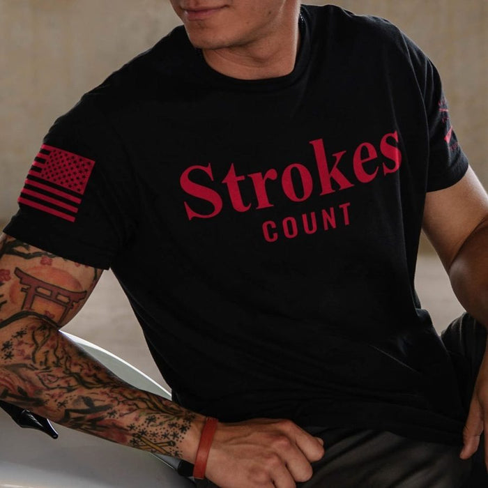 A man wearing a black short-sleeve t-shirt with 'Strokes COUNT' printed in red across the chest and a red American flag graphic on the right sleeve.