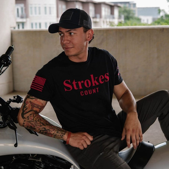 A man wearing a black t-shirt with red 'Strokes COUNT' text and a black cap sits on a white motorcycle in an outdoor setting.