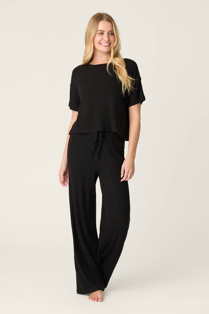 A woman with long, wavy blonde hair smiles barefoot against a plain background, wearing the P.J. Salvage Textured Essentials Black Long PJ Set featuring a cozy short-sleeve top and matching wide-leg pants.