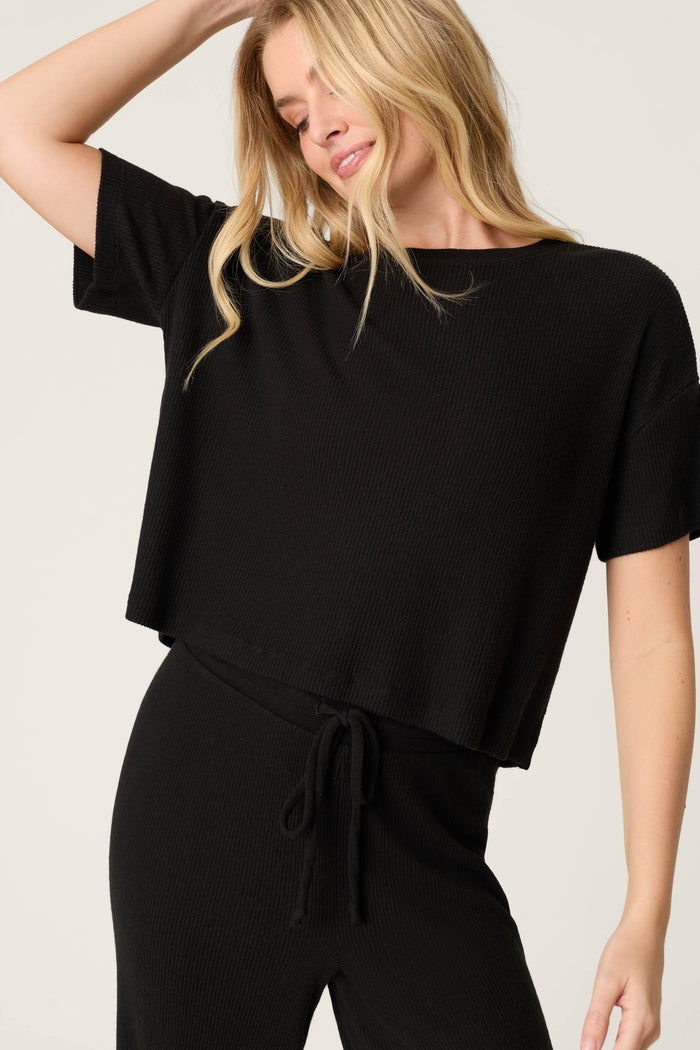 A person with long blonde hair models the P.J. Salvage Textured Essentials Black Long PJ Set, featuring a relaxed short-sleeve top and drawstring pants, smiling gently and looking down against a plain background.