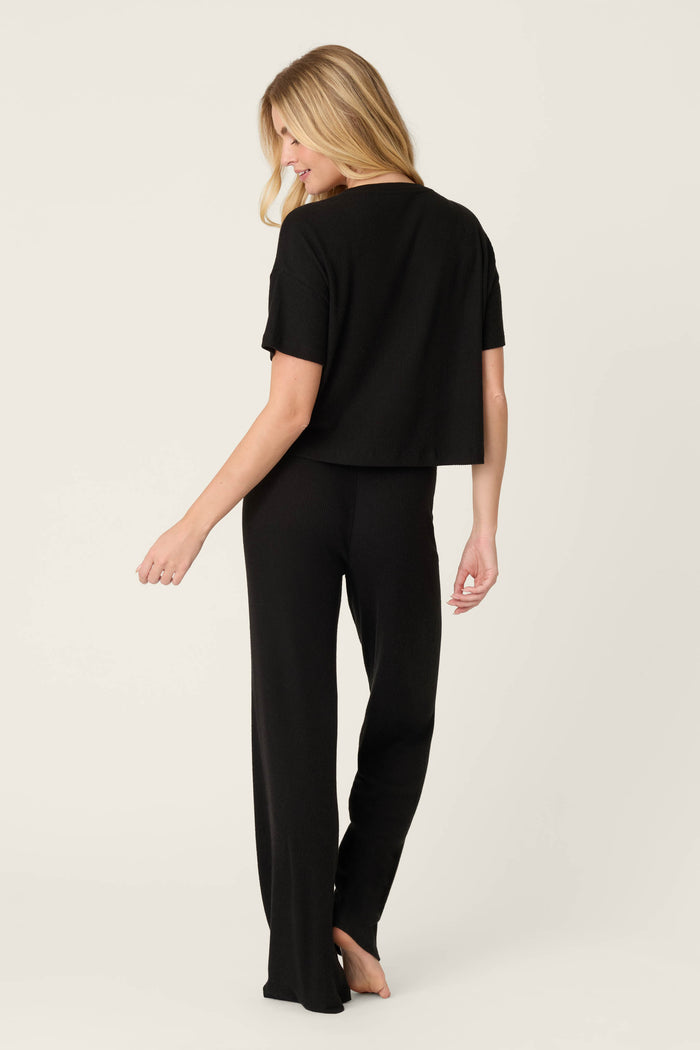A person stands barefoot, facing away from the camera, wearing the P.J. Salvage Textured Essentials Black Long PJ Set—featuring short sleeves and matching pants—against a plain light background.