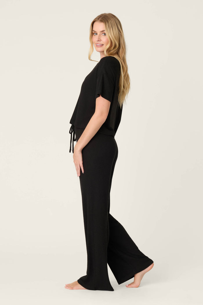 A woman stands barefoot on a plain background, sideways with one hand on her hip and a slight smile, wearing the P.J. Salvage Textured Essentials Black Long PJ Set featuring a cozy loose-fit short-sleeve top and wide-leg pants.