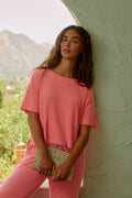 A woman with long brown hair wears the P.J. Salvage Textured Essentials Sunset Rose Short Sleeve Top and matching pants, standing against a light green textured wall with a beige patterned clutch, mountains, and lush greenery in the background.
