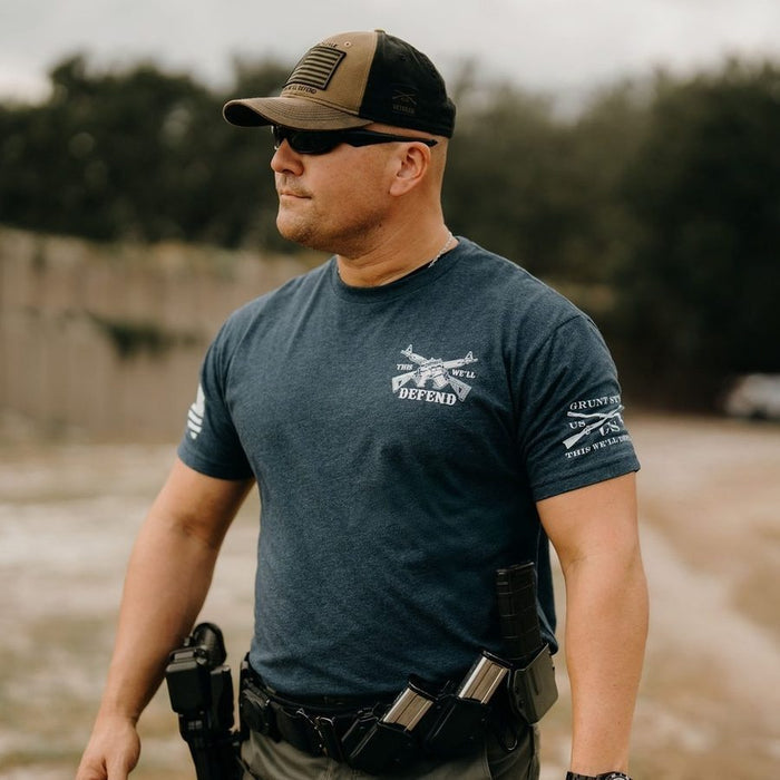 Man wearing a blue t-shirt with text and graphics, sunglasses, and a cap outdoors.