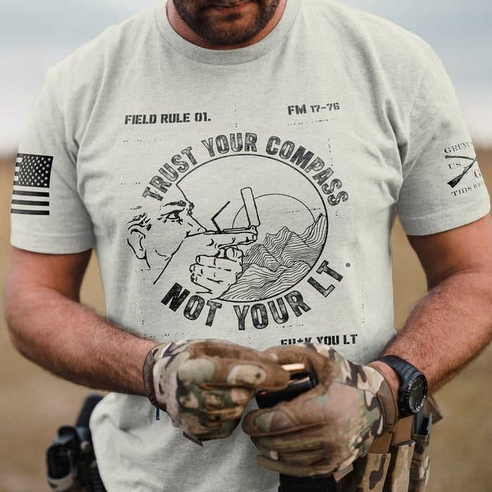 A man wearing a light gray graphic t-shirt with a black distressed print featuring a compass, rifle, and the text 'TRUST YOUR COMPASS NOT YOUR LT.' He is also wearing camouflage tactical gloves and holding a handgun.