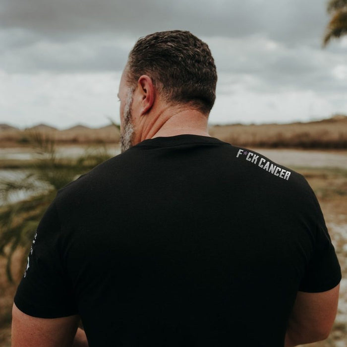 Man wearing a black t-shirt with 'F*CK CANCER' text, standing outdoors.