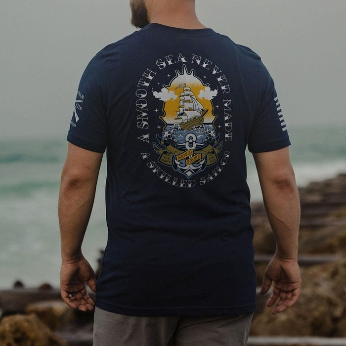 Person wearing a navy blue t-shirt with a ship graphic and text by the ocean.