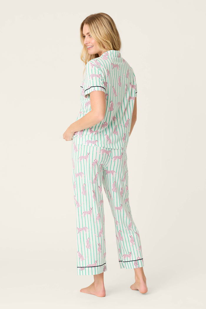 A woman stands barefoot, smiling at the camera in the P.J. Salvage Whimsy Cool Cat Cropped PJ Set—green and white stripes with pink palm tree print—against a plain off-white background.