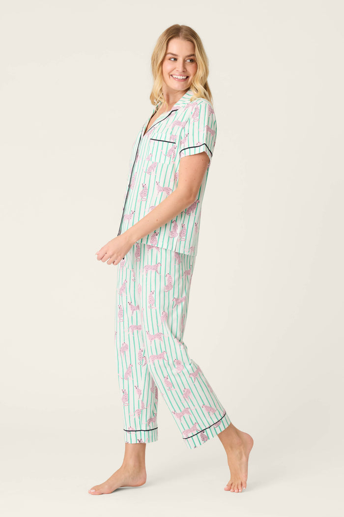 A woman stands barefoot on a light background, wearing the Whimsy Cool Cat Cropped PJ Set by P.J. Salvage, featuring a matching short-sleeve, button-front pajama top with vertical green stripes and pink flamingo patterns.
