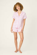 A person stands barefoot against a plain background, wearing the Whimsy Retro Shades Short PJ Set by P.J. Salvage—light pink pajamas with a small, colorful pattern, featuring a short-sleeve button-up top and matching shorts.