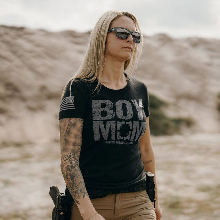 Person wearing a black t-shirt with 'BOY MOM' text, sunglasses, and holding a gun in a desert setting.