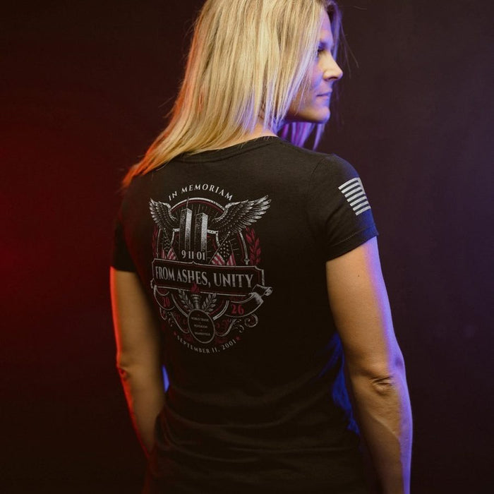 A woman shown from the back wearing a black short-sleeve graphic T-shirt with a large memorial-style print on the back and a small flag graphic on the sleeve, photographed against a dark studio background with red and blue lighting.