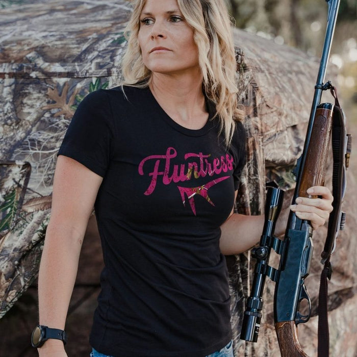 A woman wearing a black t-shirt with a pink 'Fluttress' graphic, holding a rifle in front of a camouflage hunting blind.
