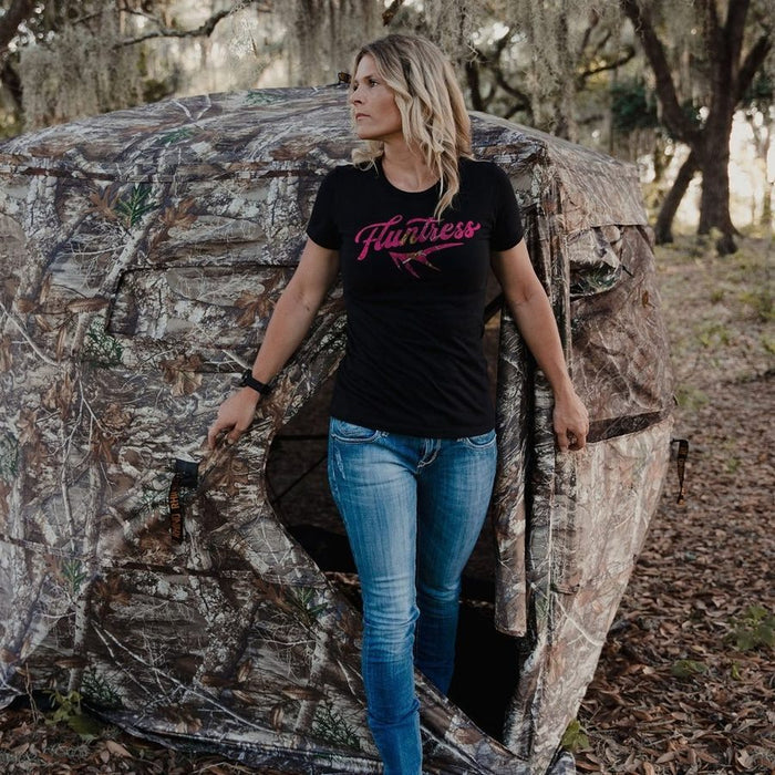 A woman wearing a black short-sleeve t-shirt with a pink 'Flintstone' graphic and blue jeans stands in the doorway of a camouflage hunting blind in an outdoor wooded setting.