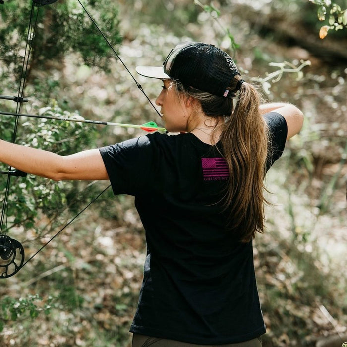 A woman in a black t-shirt and black cap aiming a compound bow in a wooded outdoor setting. The back of her shirt features a pink American flag graphic with the text 'COLUMBINE DAY' underneath.
