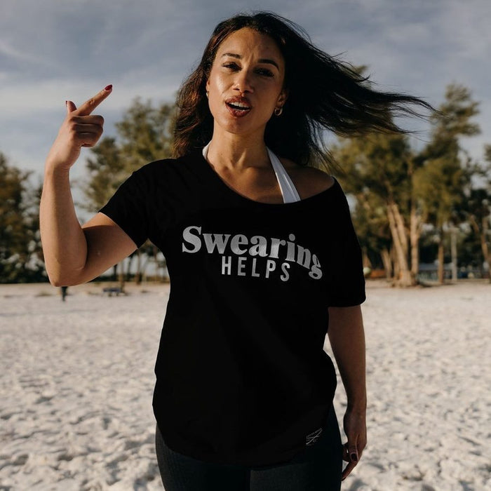 Woman wearing a black t-shirt with 'Swearing HELPS' text on a beach.