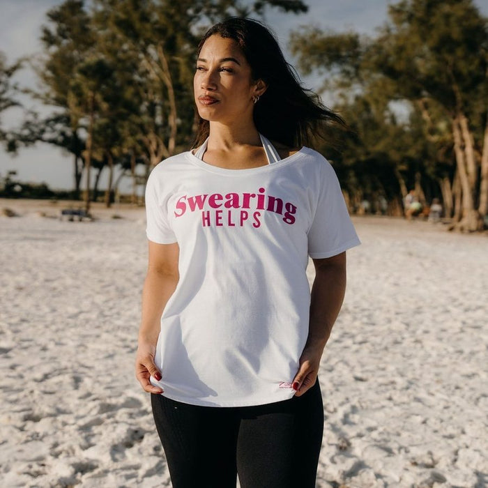 Woman wearing a white t-shirt with 'Swearing Helps' text on a sandy beach.